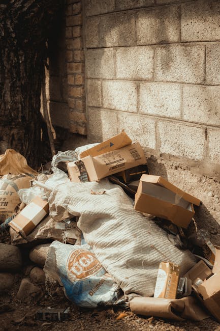 A pile of discarded cardboard boxes and paper packaging materials is situated at the base of a rough brick wall, partially sheltered by a large tree trunk on the left. The cardboard boxes vary in size, some are flattened while others retain their rectangular shape, with visible tape and printed labels on several. Intermixed with the boxes are brown paper bags and crumpled paper for packaging. The materials have a weathered appearance, with some torn or slightly crushed, indicating they have been discarded for collection or removal. The ground beneath the heap consists of small dark stones and dirt, with no visible pathways or bins nearby. The snapshot reflects a site awaiting a rubbish removal service, illustrating an instance of private waste handling or on-site clearance that might be addressed by a specialist such as Rubbish Removal Shoreditch. The setting appears outdoor and urban, with natural light casting gentle shadows and highlighting the textures of paper, cardboard, and the brick surface behind.