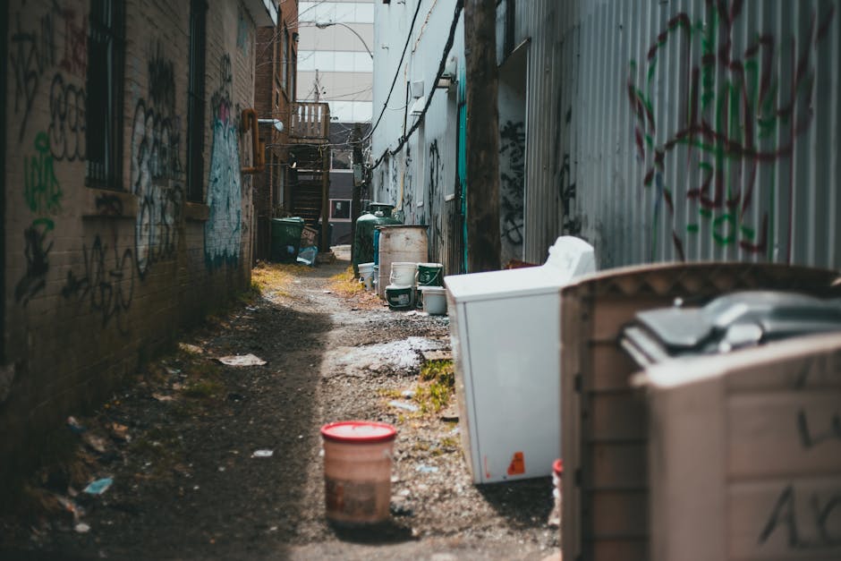 The image depicts a narrow alleyway situated between two buildings, with brick and corrugated metal walls that are heavily covered in graffiti tags and murals. The ground is uneven and muddy, with patches of standing water and scattered debris, including empty and discarded items. Several large, white plastic bags filled with waste are placed along the right side, with some partially obscured by a wooden crate or bin. Towards the background, there are green wheelie bins and a white container, likely used for waste disposal, aligned along the alley. A set of wooden stairs leading to an upper level can be seen at the far end of the alley, with attached emergency escape routes visible on the building façades. Overhead, thin black cables and wires crisscross, adding to the cluttered and neglected appearance. This scene illustrates an area that could benefit from private rubbish collection or on-site waste management, in line with services offered by Rubbish Removal Shoreditch, aimed at improving access and cleanliness in tight urban spaces.