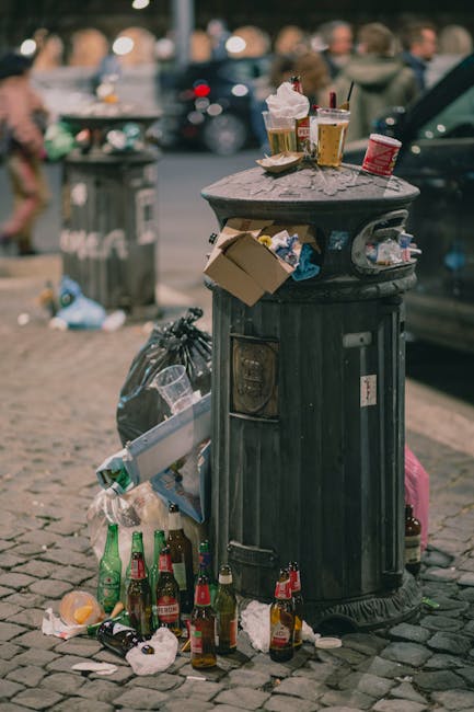 A black, vintage-style metal rubbish bin located on a cobbled street, surrounded by scattered litter including torn paper, glass bottles, and plastic bags. The top of the bin holds discarded food containers, paper cups with plastic lids, and glasses filled with beverages, some with straws. A crumpled cardboard box is partly tucked into the side of the bin, while a black trash bag leans against its side. In the background, blurred figures of pedestrians and parked cars are visible, with additional discarded waste nearby, indicative of a busy urban environment. The scene captures a moment of unmanaged litter accumulation, highlighting the importance of private waste handling and the potential need for rubbish removal services such as those offered by Rubbish Removal Shoreditch, particularly in busy areas. Lighting appears natural, with soft daylight illuminating the scene and highlighting the texture of the cobblestones and the various materials of the waste items.