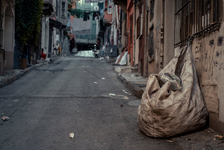 A large, weathered white trash bag filled with waste sits on an uneven sidewalk next to a building with a rough, graffiti-marked wall in an urban alleyway. The bag appears to contain mixed rubbish, with some plastic packaging visible at the top. In the background, the narrow street slopes gently upward, lined with closely packed residential and commercial buildings constructed from various materials, including brick and concrete. Several minor pieces of litter are scattered on the pavement, and laundry or blankets are hanging on clotheslines strung between buildings in the distance. The lighting suggests an overcast day, with diffused natural light illuminating the scene. The area looks cluttered and in need of cleaning, illustrating an environment where independent rubbish removal services, such as those provided by Rubbish Removal Shoreditch, might be beneficial for on-site clearance or alternative waste handling solutions in busy urban settings.