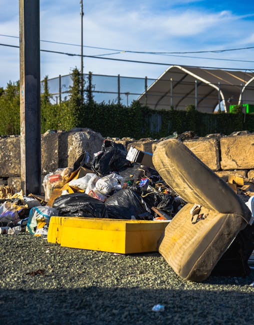 A black, vintage-style metal rubbish bin located on a cobbled street, surrounded by scattered litter including torn paper, glass bottles, and plastic bags. The top of the bin holds discarded food containers, paper cups with plastic lids, and glasses filled with beverages, some with straws. A crumpled cardboard box is partly tucked into the side of the bin, while a black trash bag leans against its side. In the background, blurred figures of pedestrians and parked cars are visible, with additional discarded waste nearby, indicative of a busy urban environment. The scene captures a moment of unmanaged litter accumulation, highlighting the importance of private waste handling and the potential need for rubbish removal services such as those offered by Rubbish Removal Shoreditch, particularly in busy areas. Lighting appears natural, with soft daylight illuminating the scene and highlighting the texture of the cobblestones and the various materials of the waste items.